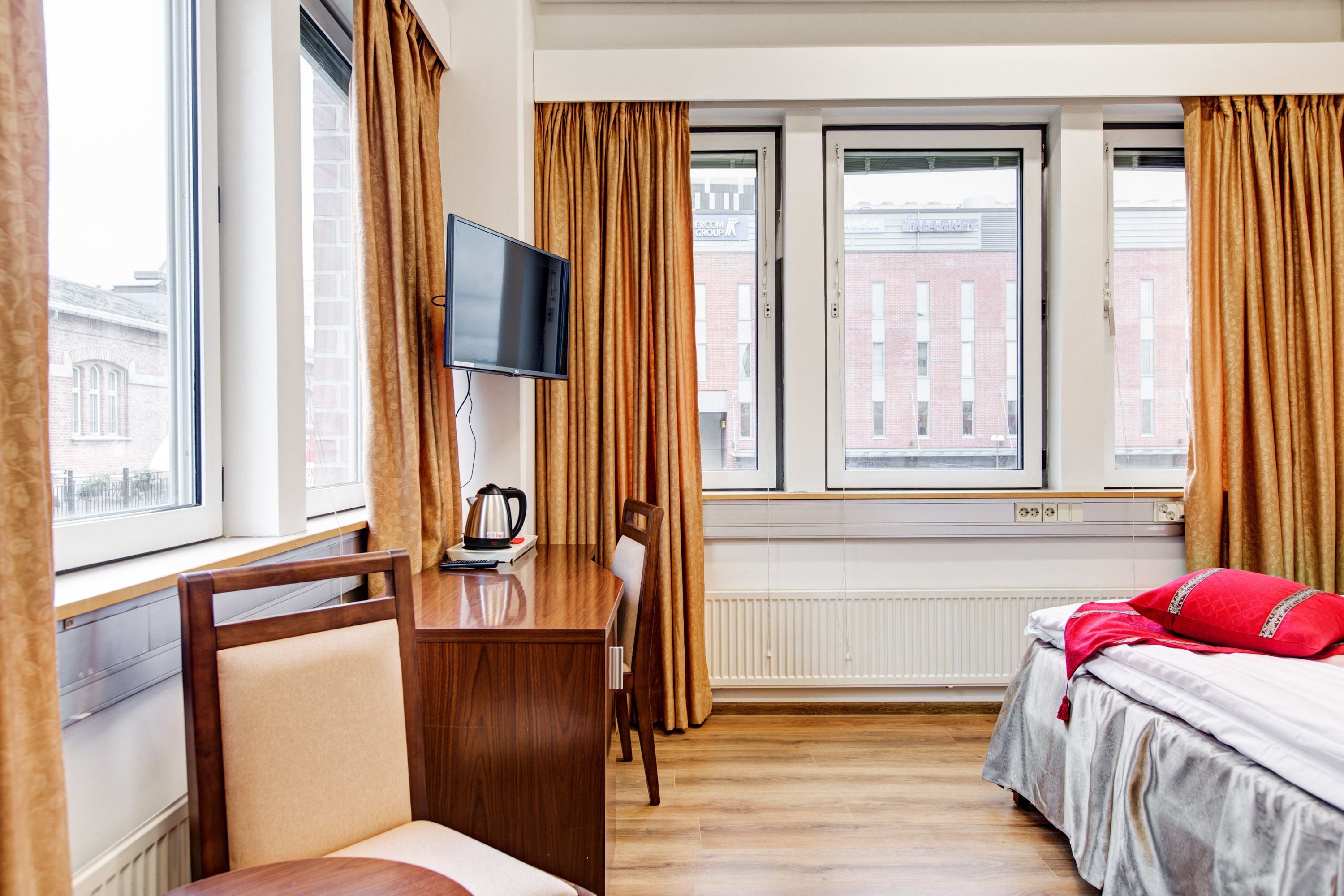 A hotel room with a bed, a desk with a chair, a television, and large windows with curtains.