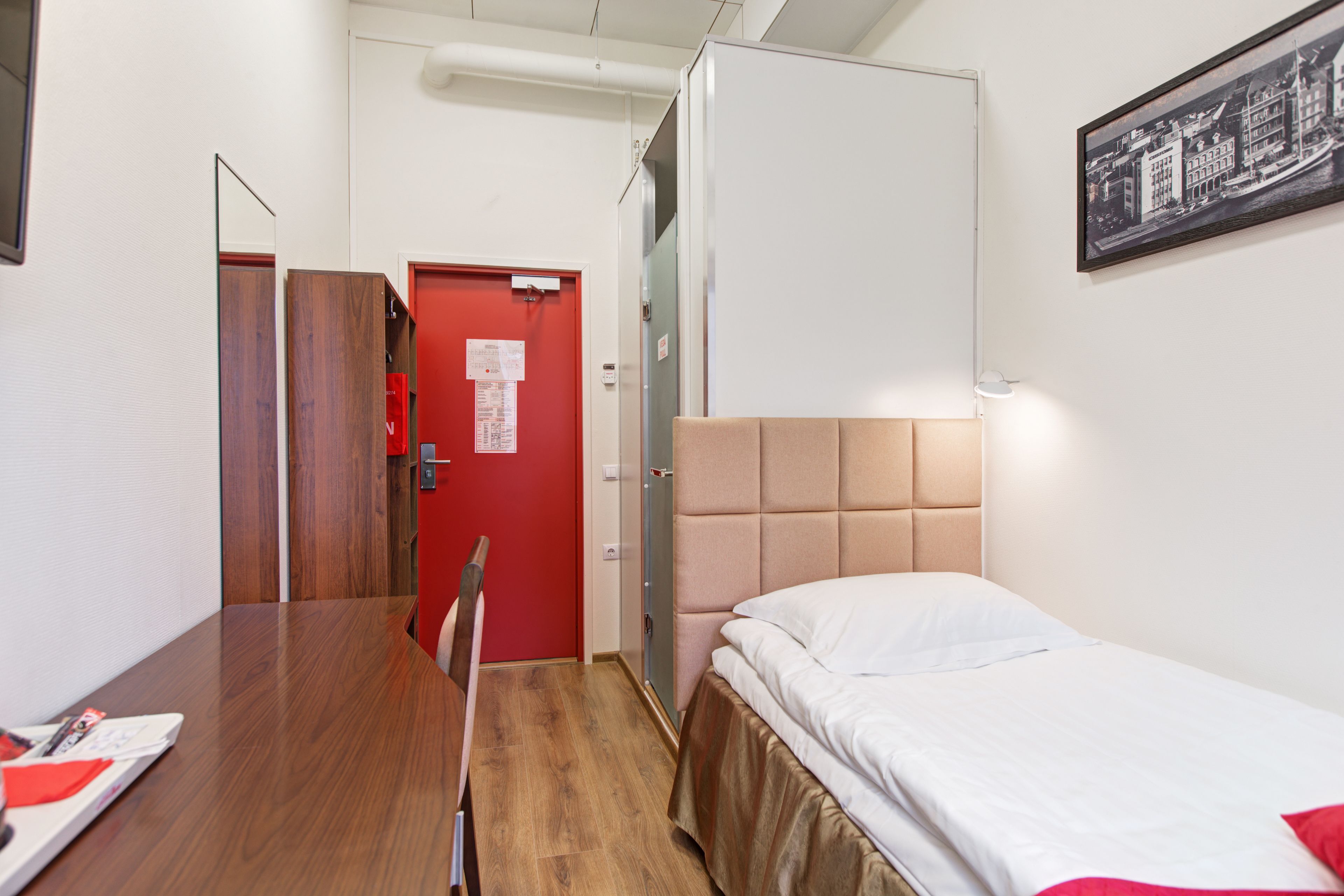 A hotel room with a wooden table, a bed with a beige headboard, a red door, and a framed picture on the wall.