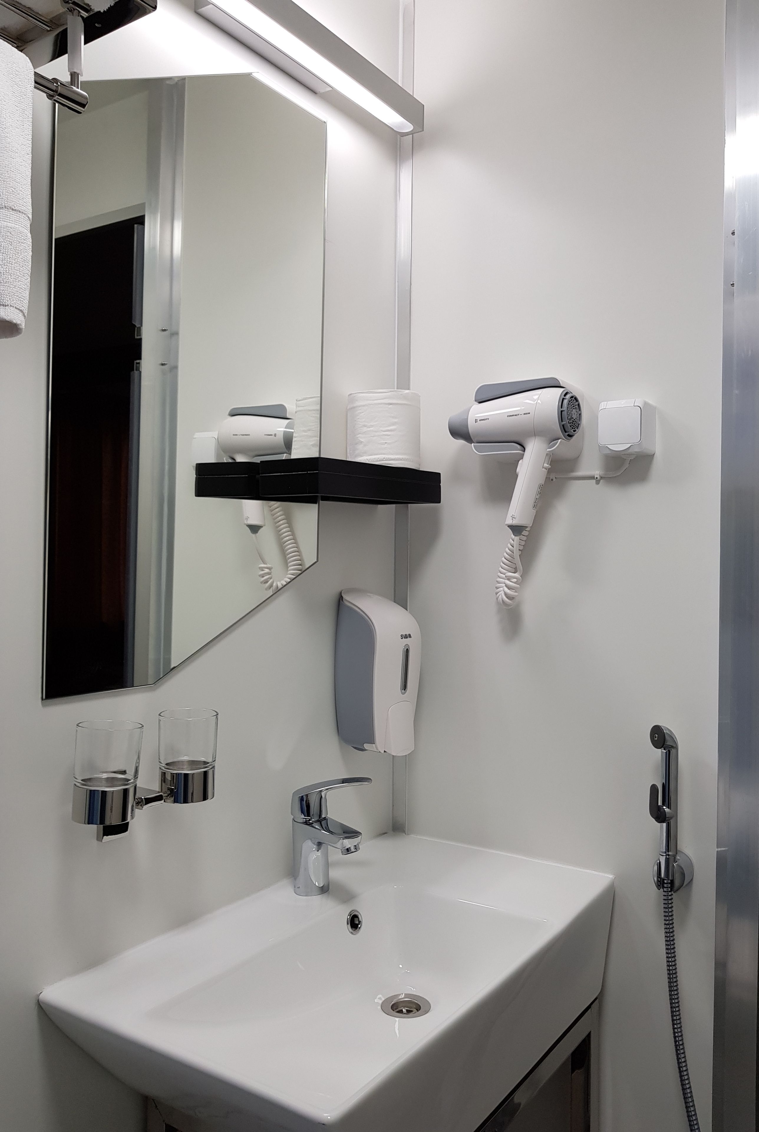 A bathroom with a white sink, a mirror, a hairdryer, a towel rack, and a soap dispenser.