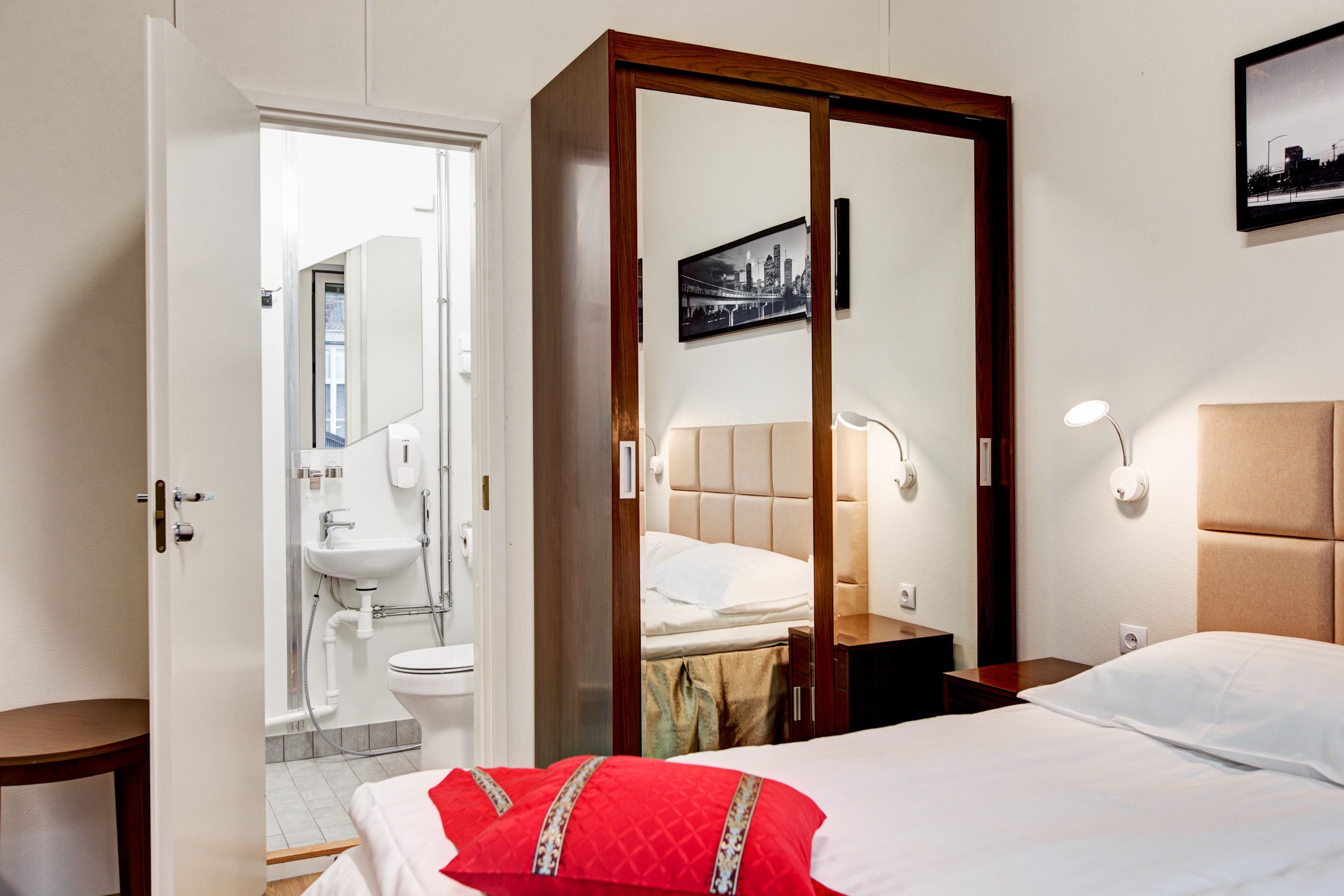 A hotel room with a bed, a wardrobe, a bedside table, a nightstand, a mirror, and a bathroom visible through an open door.