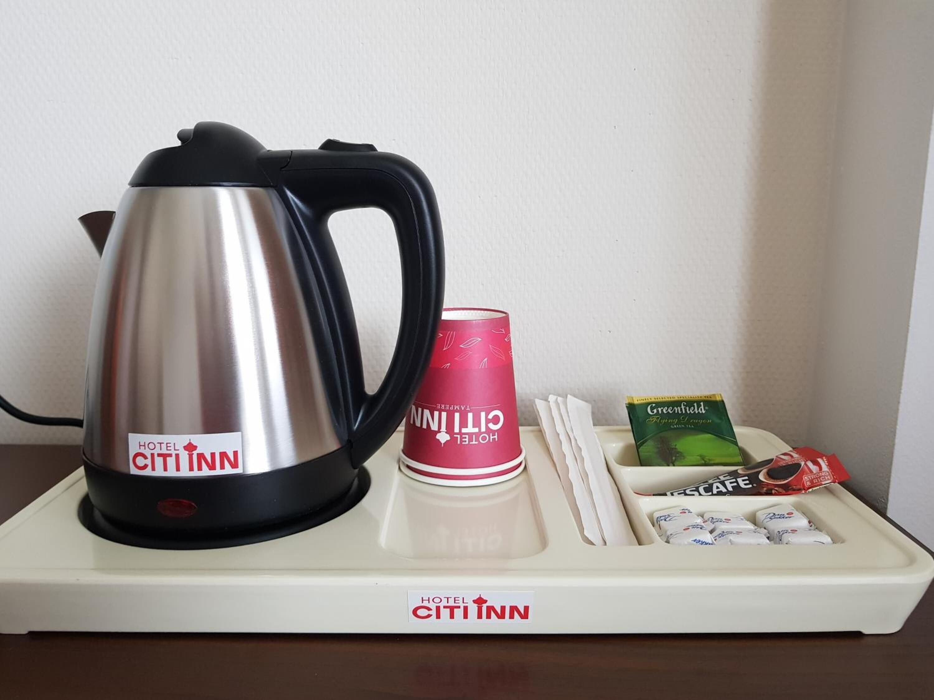 A hotel minibar setup with a kettle, a red cup, a green tea box, and a tray with snacks.