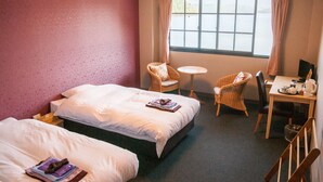 Habitación de hotel con dos camas, una mesita redonda pequeña, dos sillones de mimbre, un escritorio con computadora y tetera, y una ventana con cortinas.