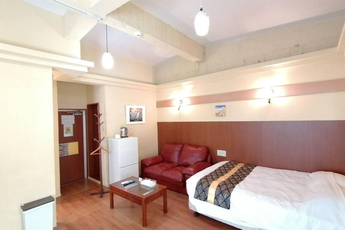 A hotel room with a bed, a red leather sofa, a small wooden table, a white radiator, and a wall-mounted light fixture.