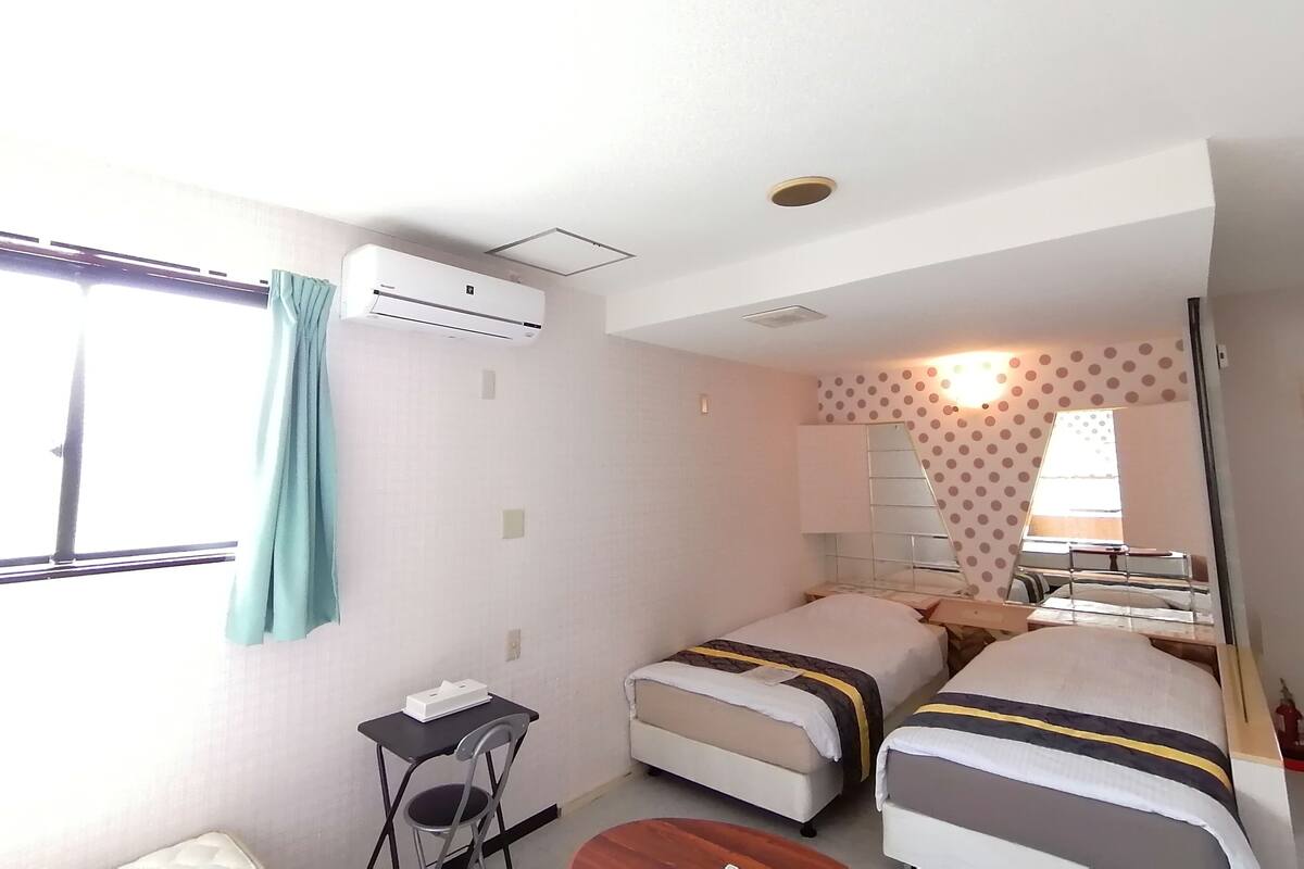 A hotel room with two beds, a desk, a chair, an air conditioning unit, and a window with curtains.