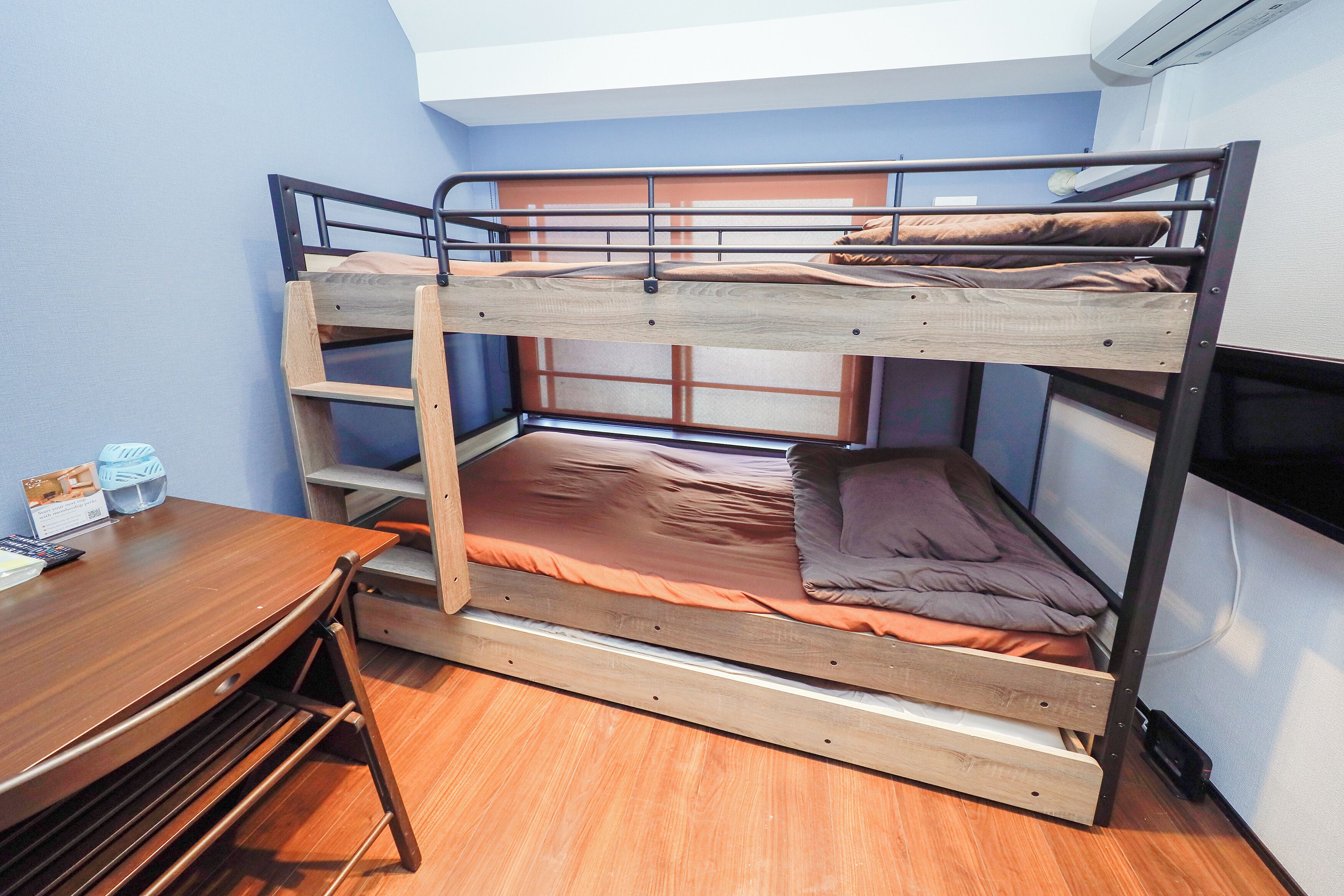 A bunk bed with a desk and chair, a small table with items, and a wall-mounted air conditioner.