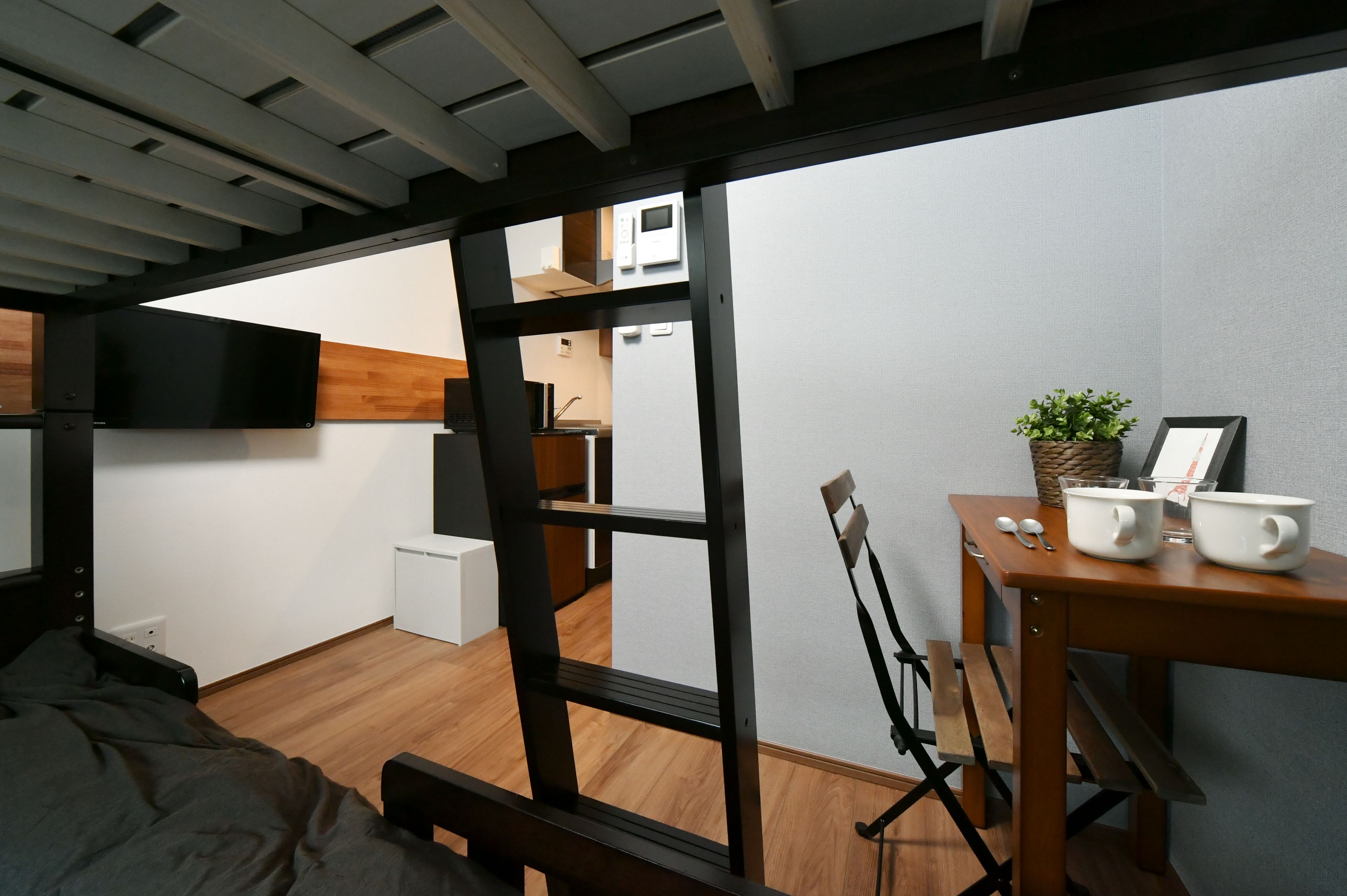 Habitación pequeña con cama, una mesa de madera con tazas y una planta, y una kitchenette al fondo.