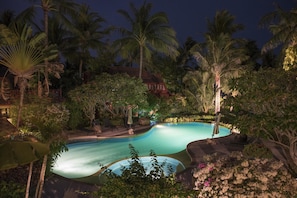 A nighttime view of a resort pool surrounded by palm trees and lush vegetation.