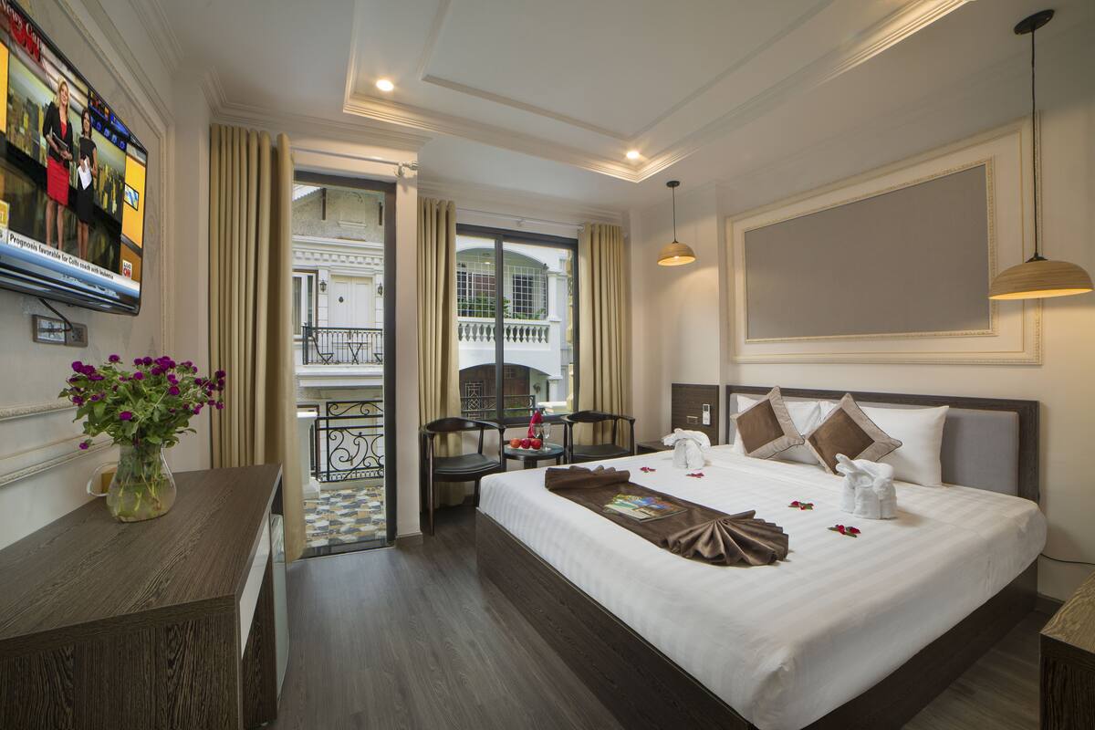 Une chambre d’hôtel avec un grand lit, un bureau agrémenté d’un vase de fleurs, une télévision et un balcon avec des rideaux.