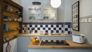Una cocina con estantes de madera, un armario de vidrio, salpicadero de azulejos, un frutero, un tostador y una lámpara colgante.