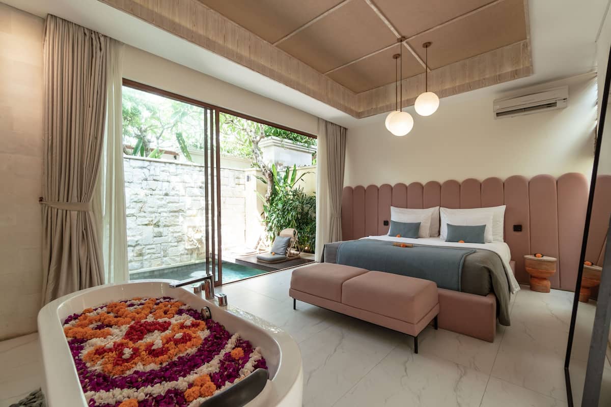 A modern bedroom with a large bed, a small sofa, a bathtub with flower petals, and a view of a garden through the sliding doors.