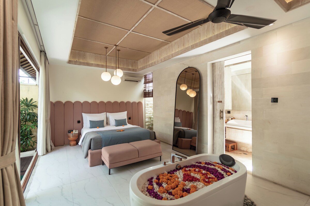 A modern bedroom with a large bed, a sitting area, a bathtub filled with flowers, and a large mirror.
