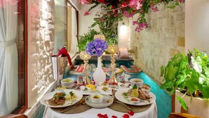 A table set with various dishes, a vase of purple flowers, and a candle on a balcony with a stone wall and greenery. - La Vie Villa Seminyak by Ini Vie Hospitality (Legian)