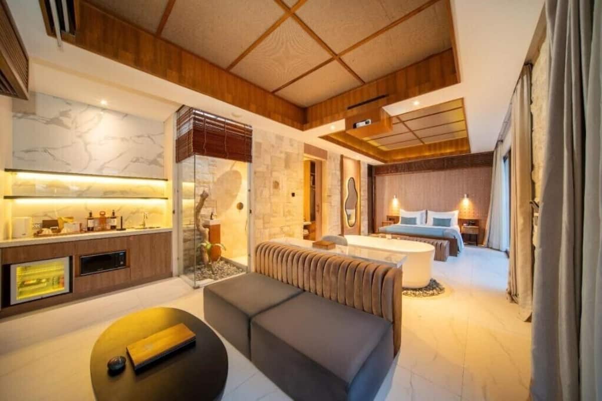 A modern hotel room with a wooden ceiling, a marble accent wall, a sofa, a dining area, and a bed with a view.