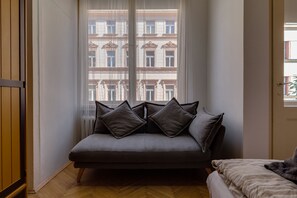 A corner room with a grey sofa, two pillows, a wooden floor, and a window with a view of buildings.