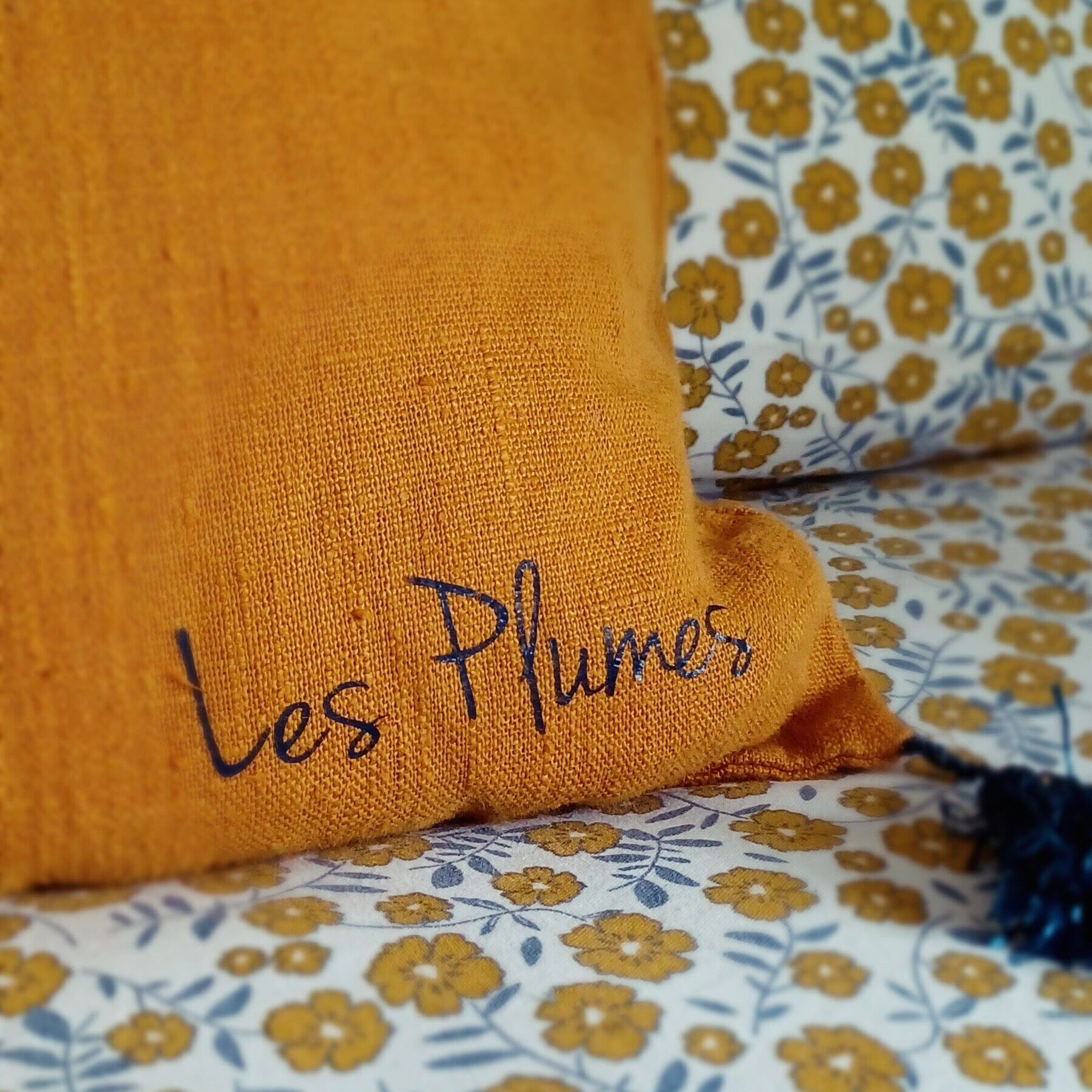Almohada amarilla con bordado que dice "Les Plumes" sobre una colcha de estampado floral.