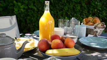 A breakfast table with a teapot, a bottle of orange juice, a bowl of fruit, and a basket of bread.