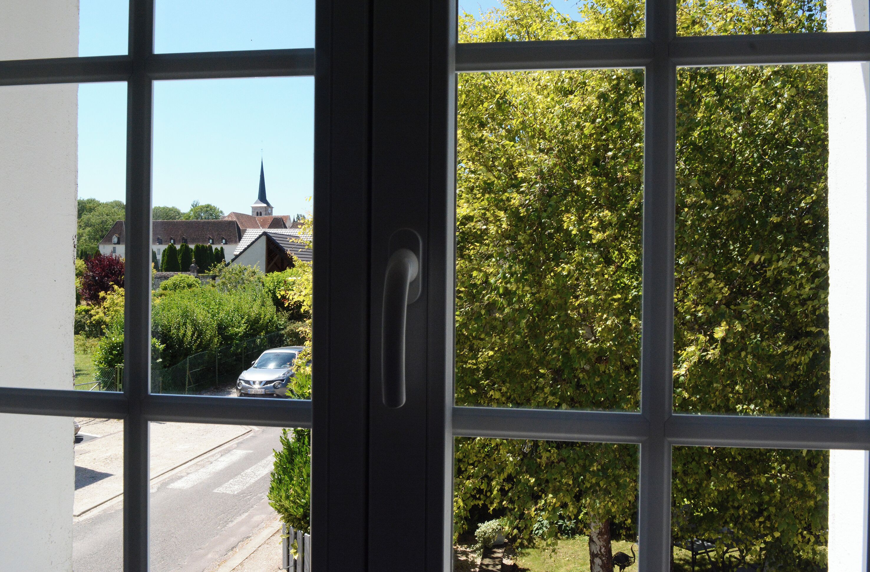 Une vue à travers une fenêtre dont le cadre est en métal, donnant sur une rue et un bâtiment surmonté d’une flèche, visible au loin.