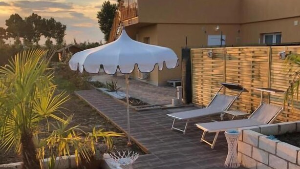 Une terrasse en bois avec des chaises longues et un parasol blanc, entourée de plantes et d’un mur de pierre.