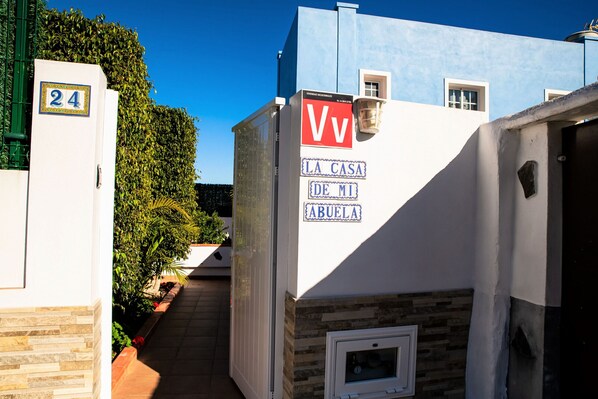 A narrow pathway leading to a building with a blue and white facade, a sign reading "VV LA CASA DE MI ABUELA", and a number "24" on a white pillar. - La casa de mi abuela with heatable pool (Garachico)