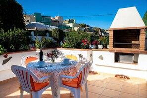 Patio esterno con tavolo apparecchiato per mangiare, circondato da piante in vaso e un barbecue.
