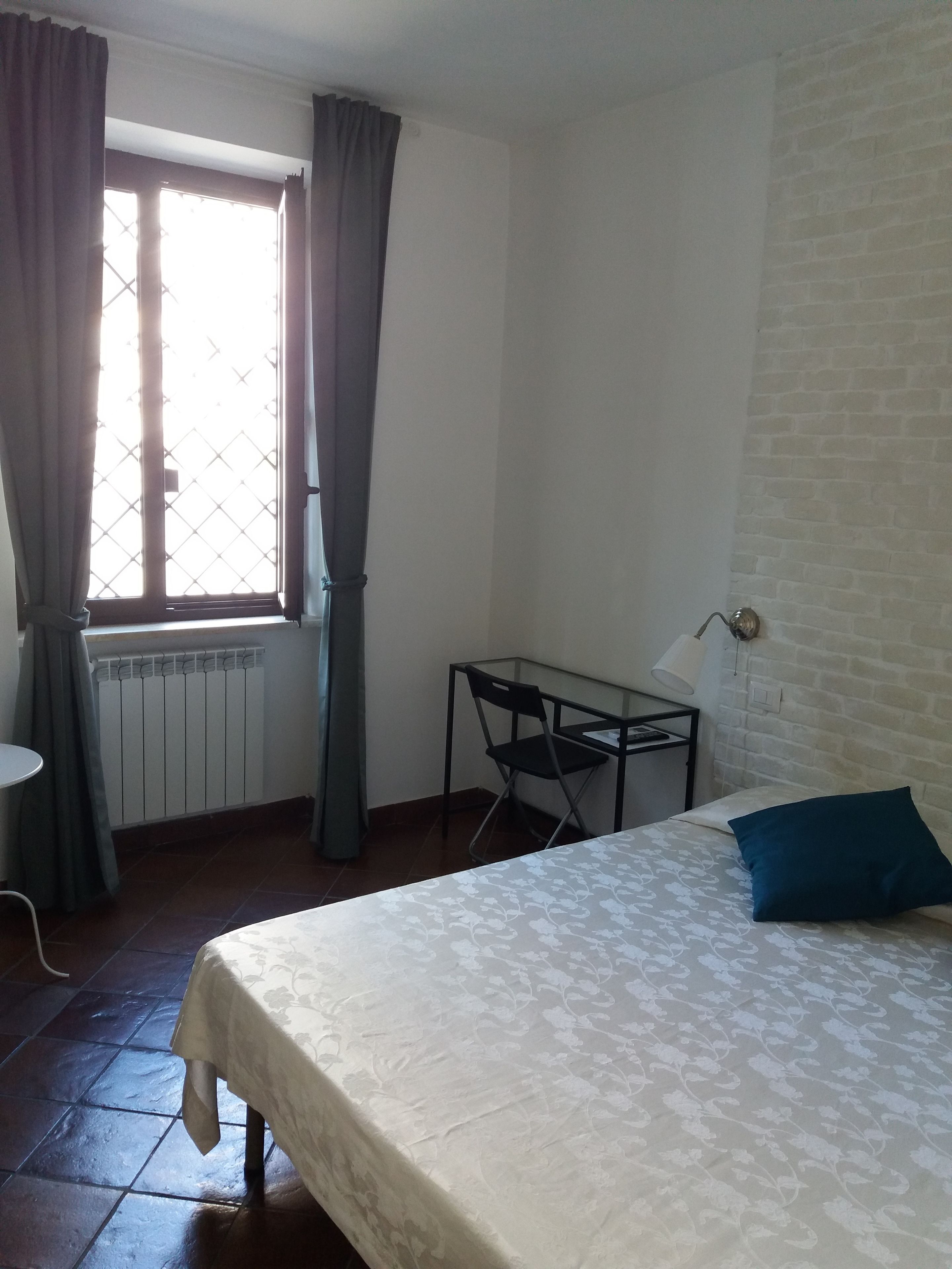 A room with a bed, a table, a chair, a window with curtains, and a radiator.