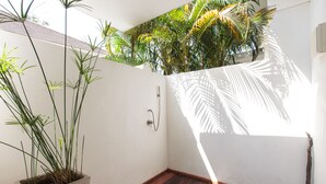 Un espacio de ducha pequeño y cerrado con piso de madera, una maceta de concreto con plantas altas y vistas a palmeras a través de una ventana.