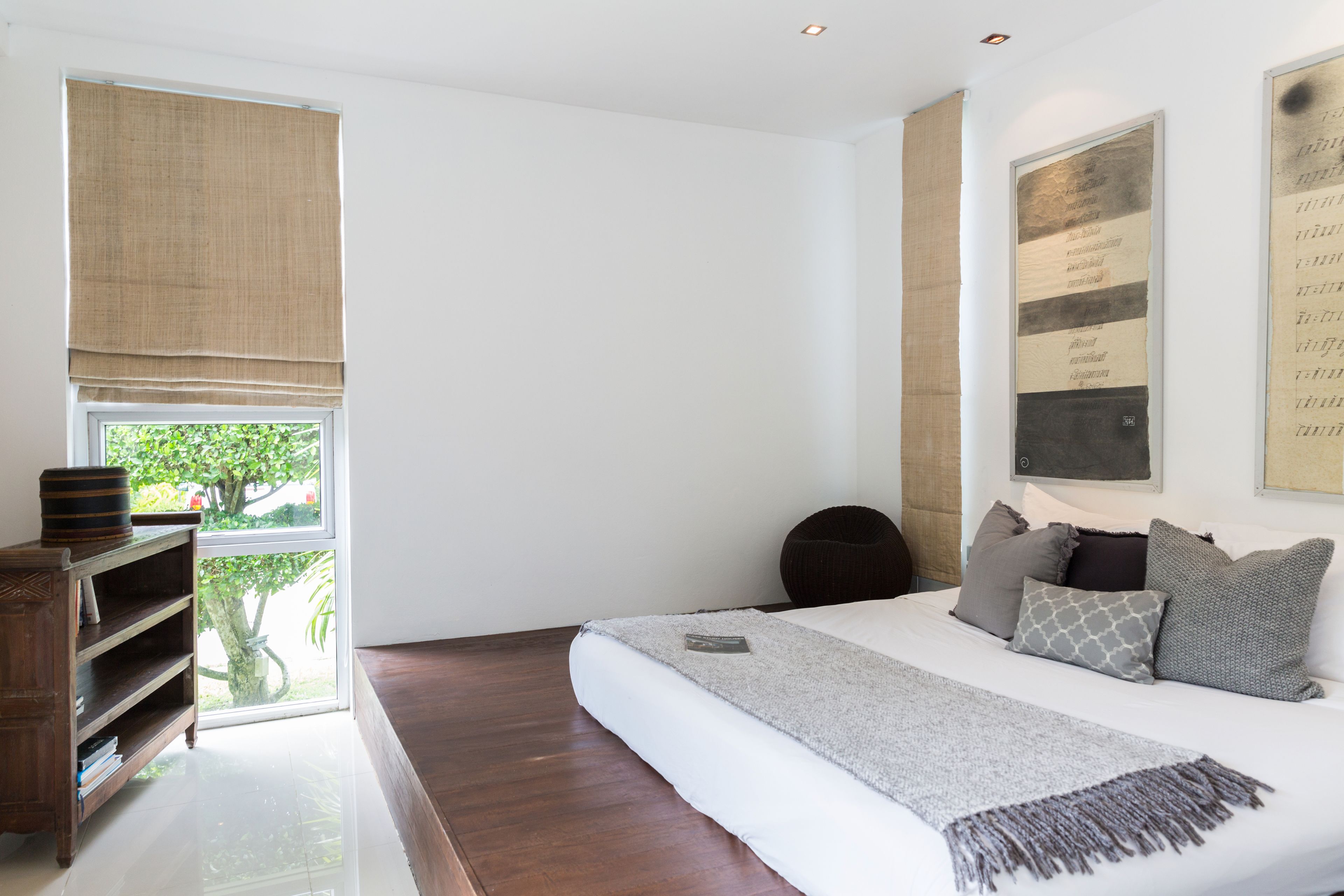 Une chambre avec un grand lit, une table de chevet en bois, une fenêtre donnant sur un paysage verdoyant et une œuvre d’art accrochée au mur.