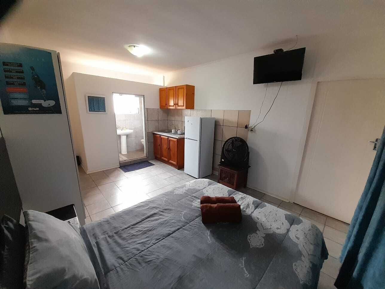 A compact room with a bed, a small kitchenette, a TV mounted on the wall, and a door leading to another room.