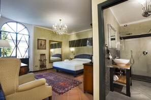 A bedroom with a large bed, a sitting area, a chandelier, a large window, and a bathroom with a sink and shower. - Corte Rosantico (Bussolengo)