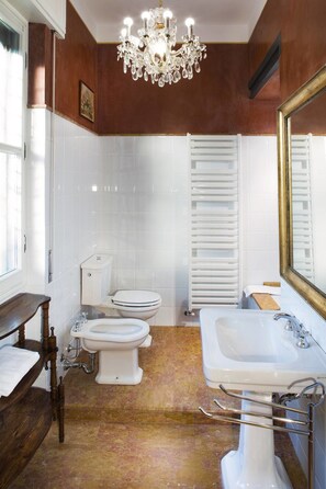 A bathroom with a chandelier, two toilets, a sink, and a wooden bench. - Corte Rosantico (Bussolengo)