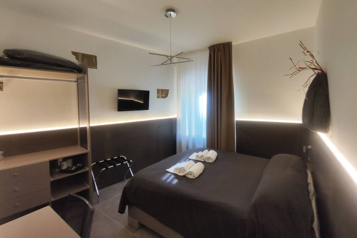 Quarto de hotel moderno com cama, escrivaninha, cadeira, televisão de tela plana e janela com cortinas.