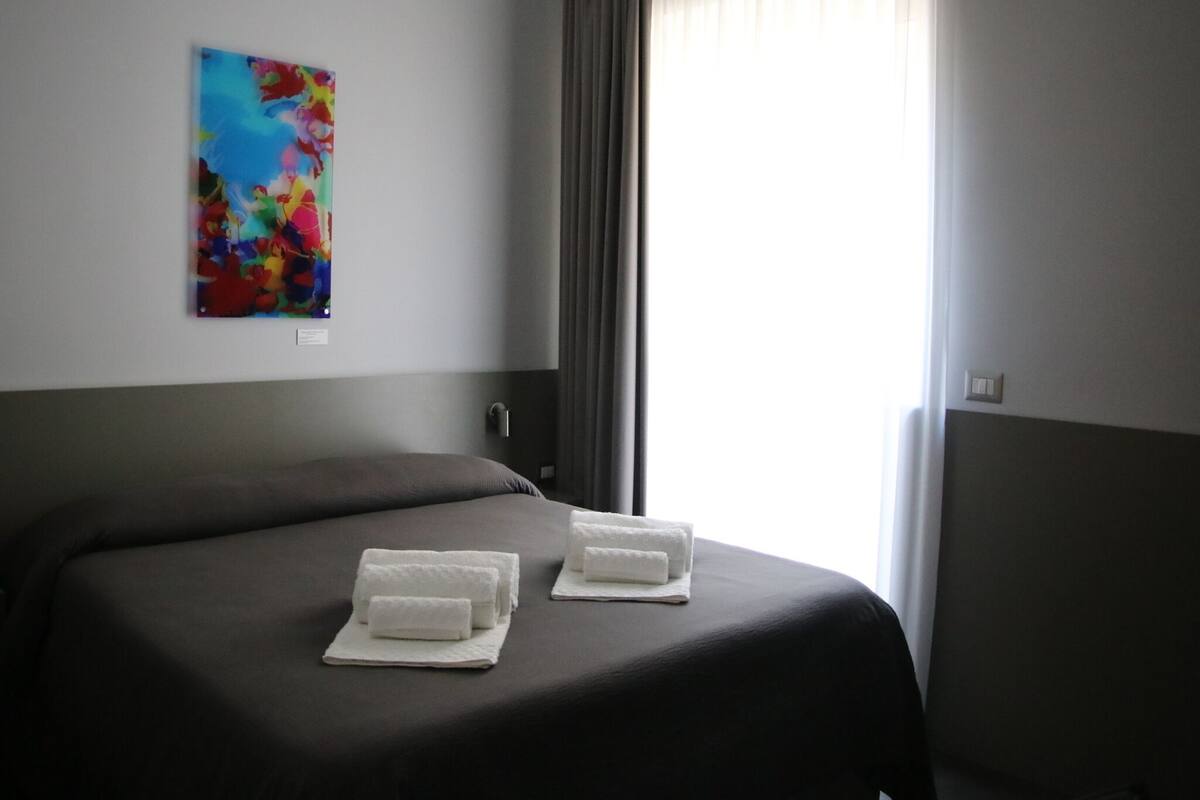 Quarto de hotel com uma cama grande, duas toalhas brancas, um quadro abstrato colorido na parede e uma janela com cortinas.