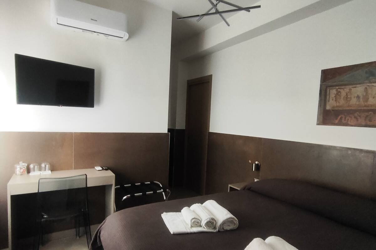 Quarto de hotel com cama, escrivaninha com cadeira, TV de tela plana na parede e ar condicionado.
