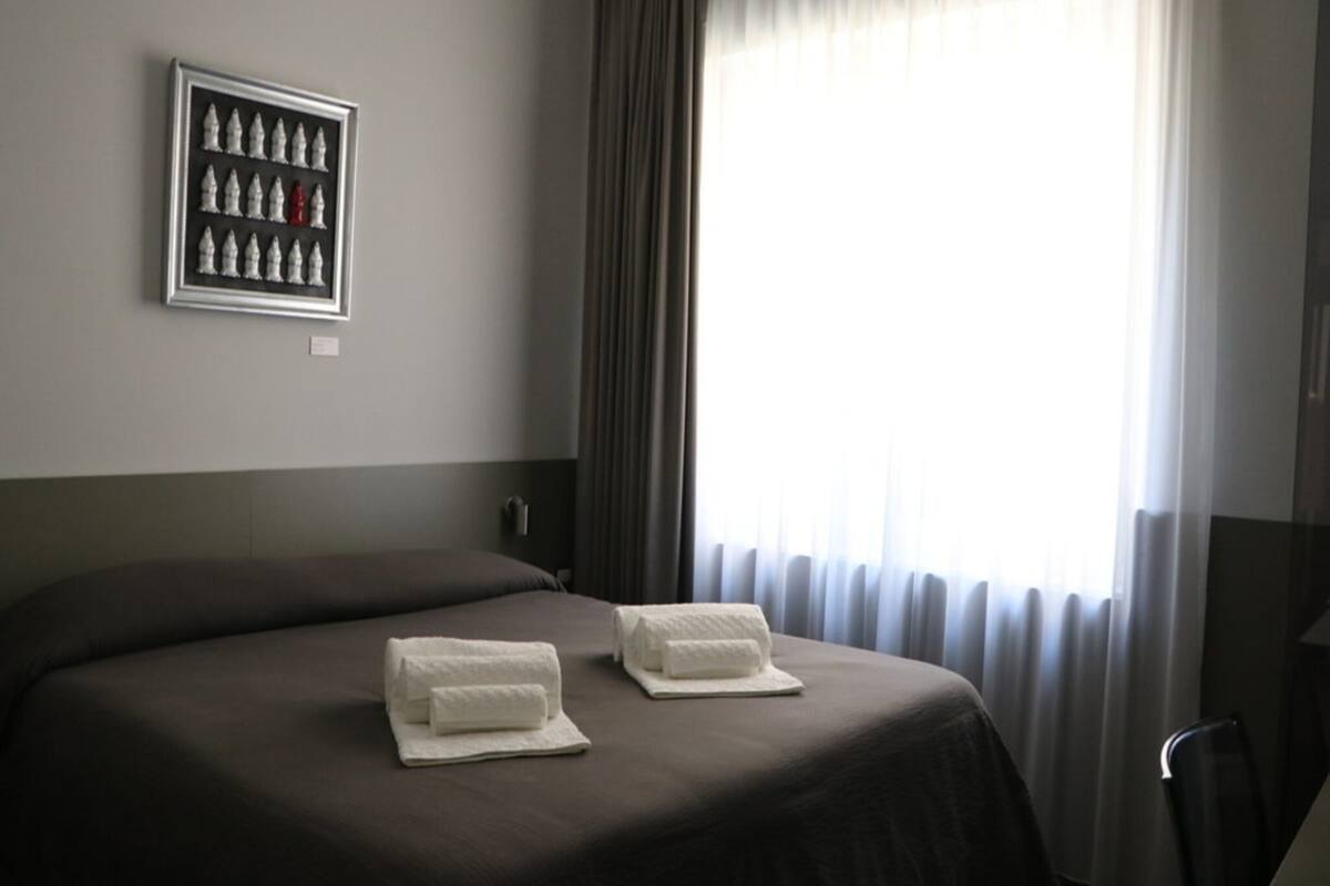 Quarto de hotel com uma cama grande, um tabuleiro de xadrez emoldurado na parede e uma janela com cortinas.