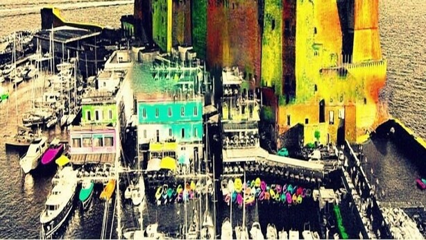 Un pueblo costero con una imponente estructura similar a un castillo, un puerto deportivo con barcos y un muelle con sombrillas de colores.
