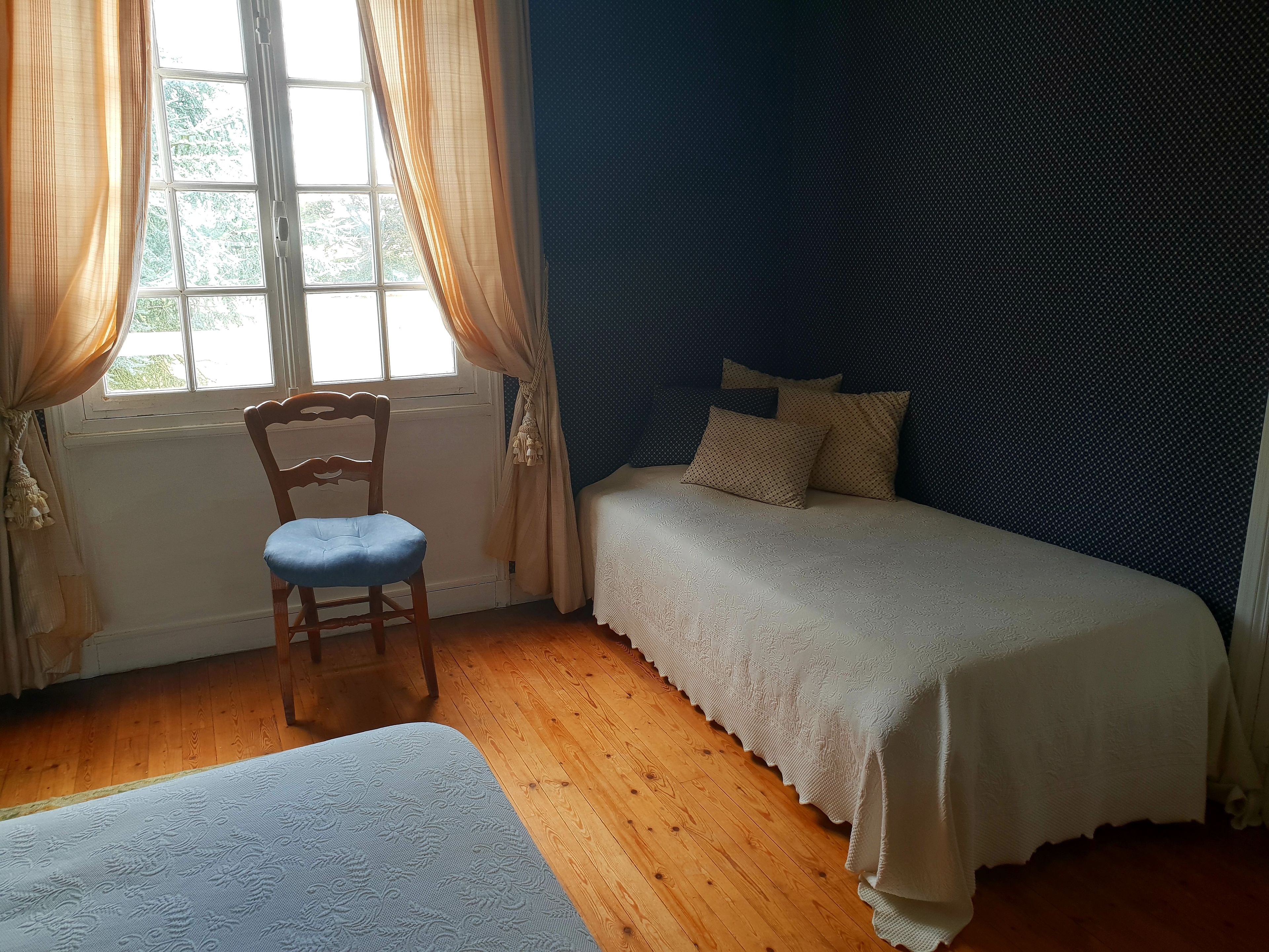 A bedroom with a wooden floor, a bed with a white bedspread, a chair with a blue cushion, and a window with curtains.