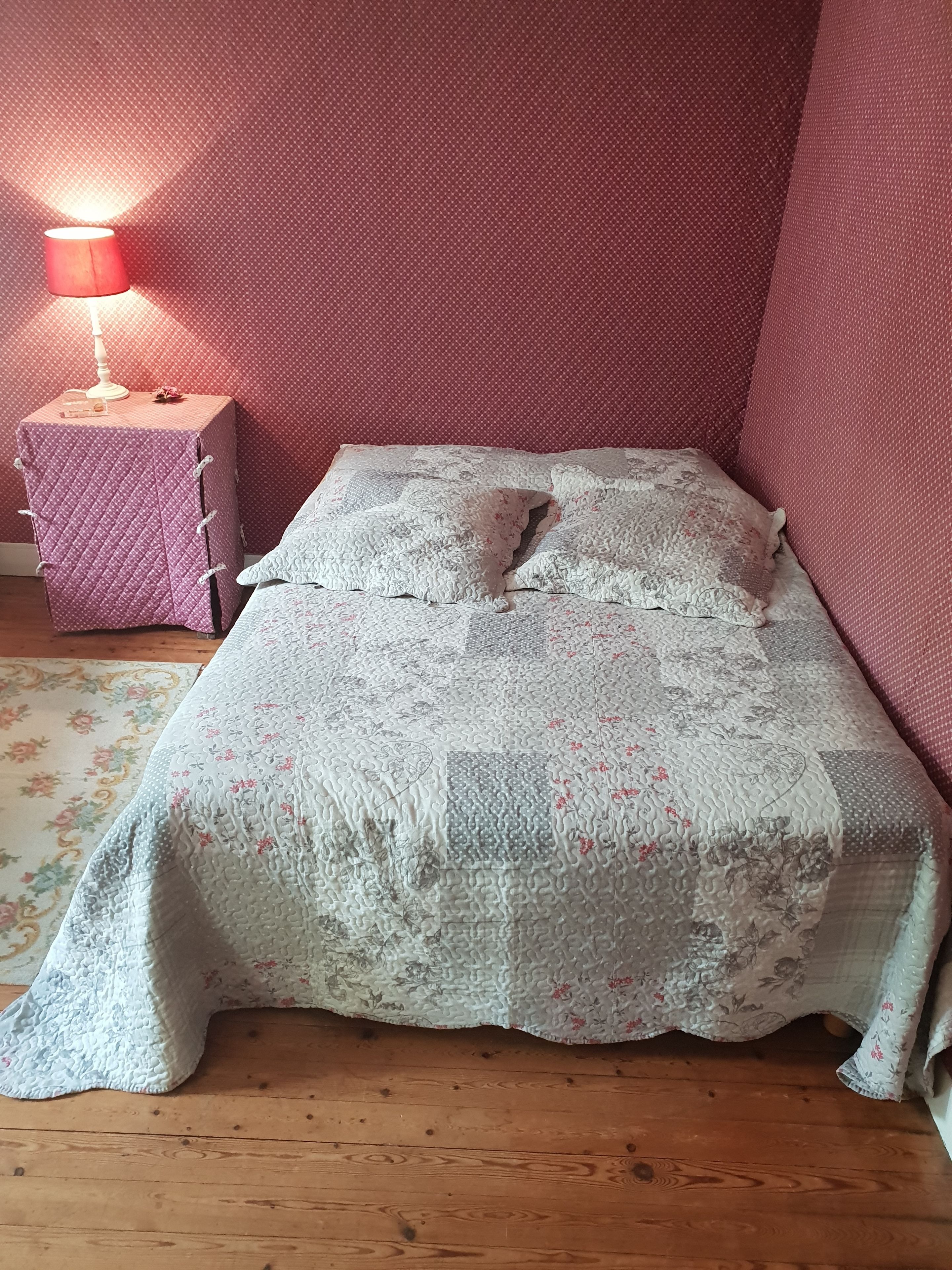 Ein kleines, gemütliches Schlafzimmer mit Holzboden, einer zudeckenden Bettdecke mit Blumenmuster und einem kleinen Nachttischchen mit Lampe.