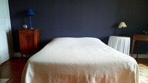 A neatly made bed with a white bedspread, a wooden nightstand with a blue lamp, and a patterned wall in the background. - Domaine de la Coudraye (La Haye-du-Theil)