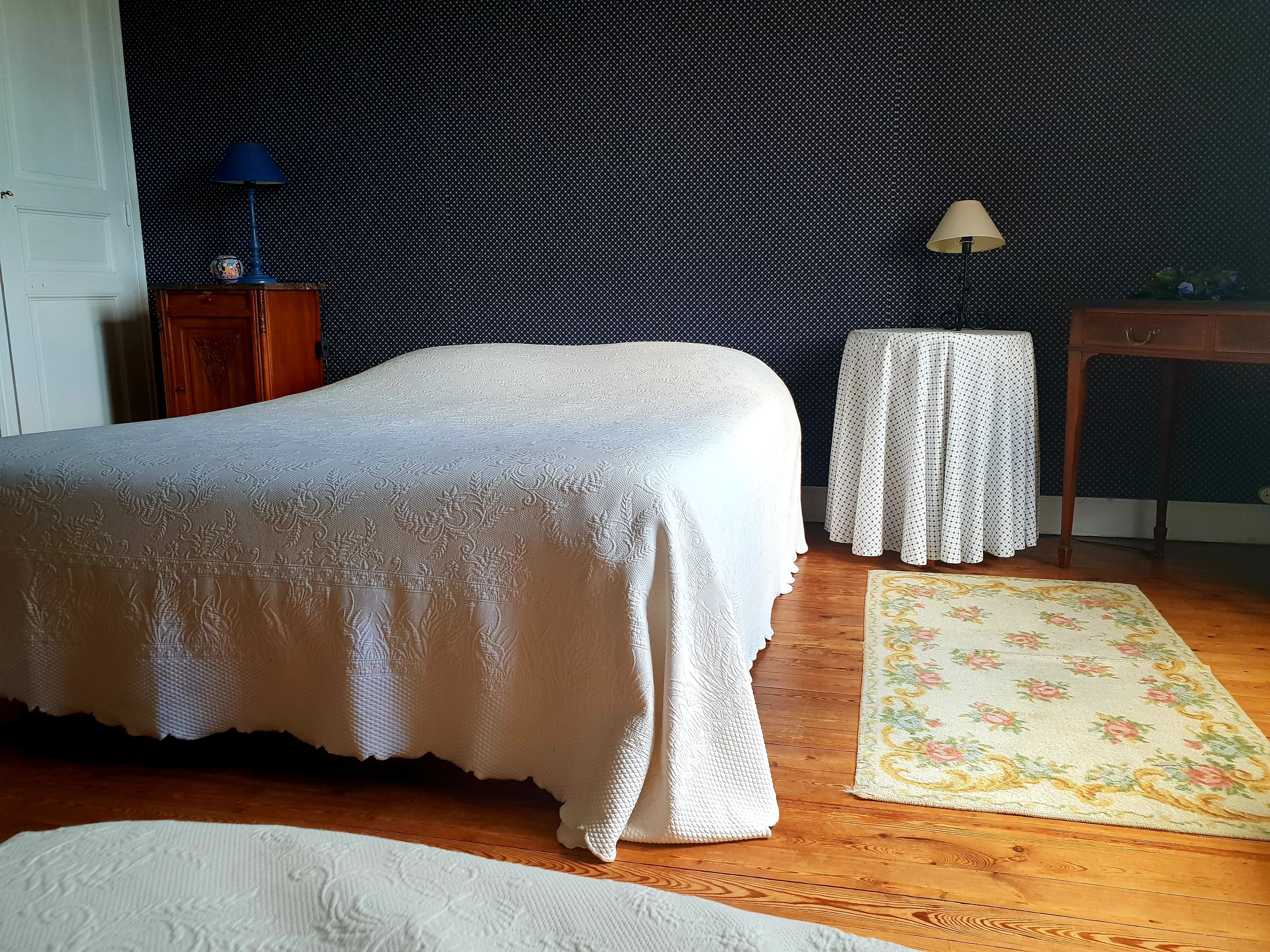 Un dormitorio ordenado con cama, una mesita, una lámpara y un tapete floral.