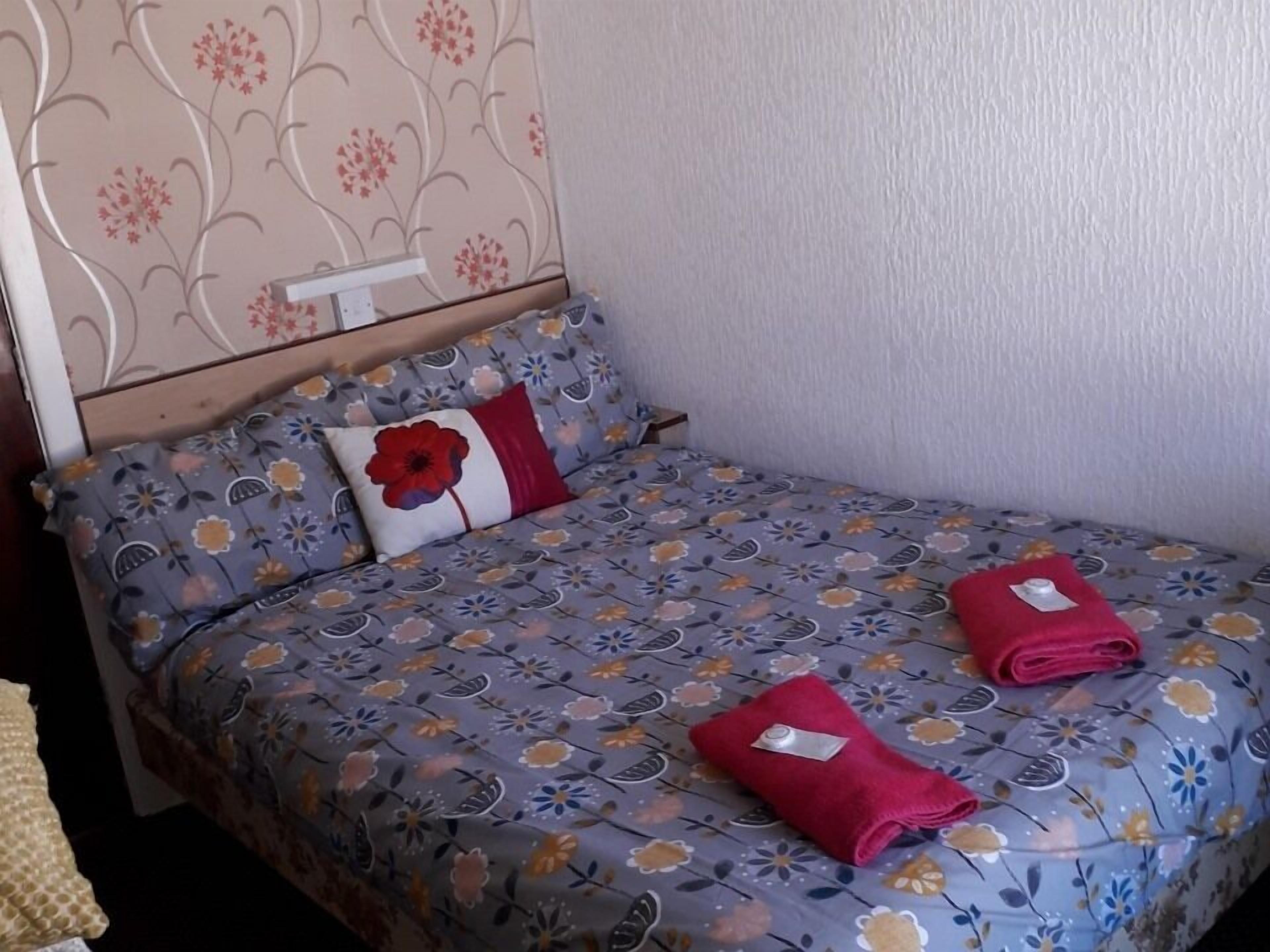 Een net opgemaakt bed met bloemenbeddengoed, een rood kussen en twee gevouwen handdoeken op het bed.