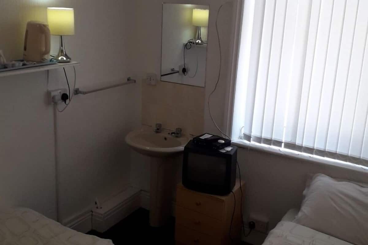 Quarto de hotel pequeno e com pouca luz, com uma cama de solteiro, uma mesa de cabeceira, um pequeno armário e uma pia com espelho.