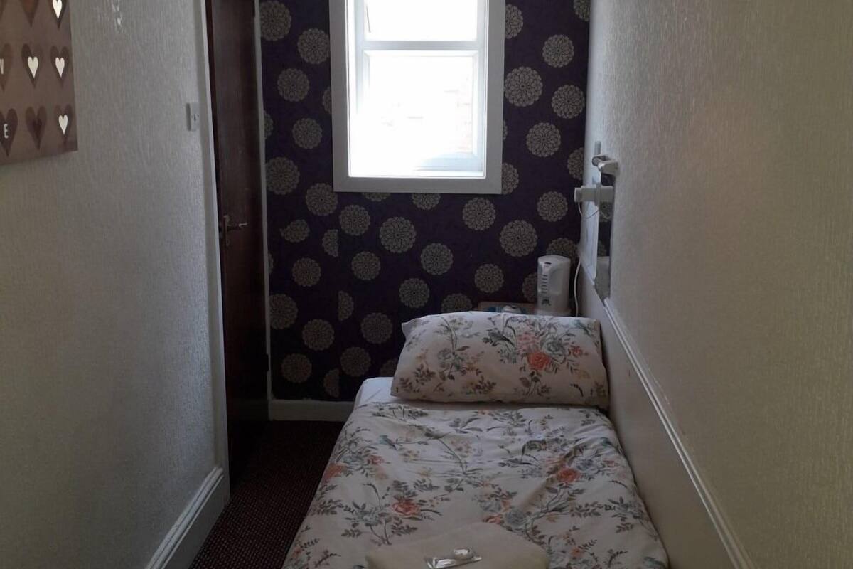 Um quarto pequeno com uma cama de solteiro, roupa de cama floral, uma janela com vista e parede estampada.