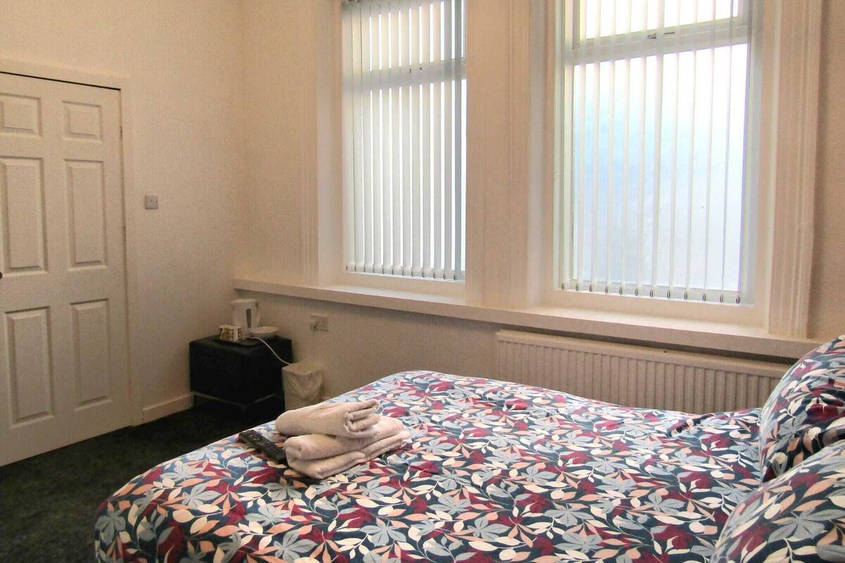 Quarto com cama, duas janelas com persianas, radiador e porta.