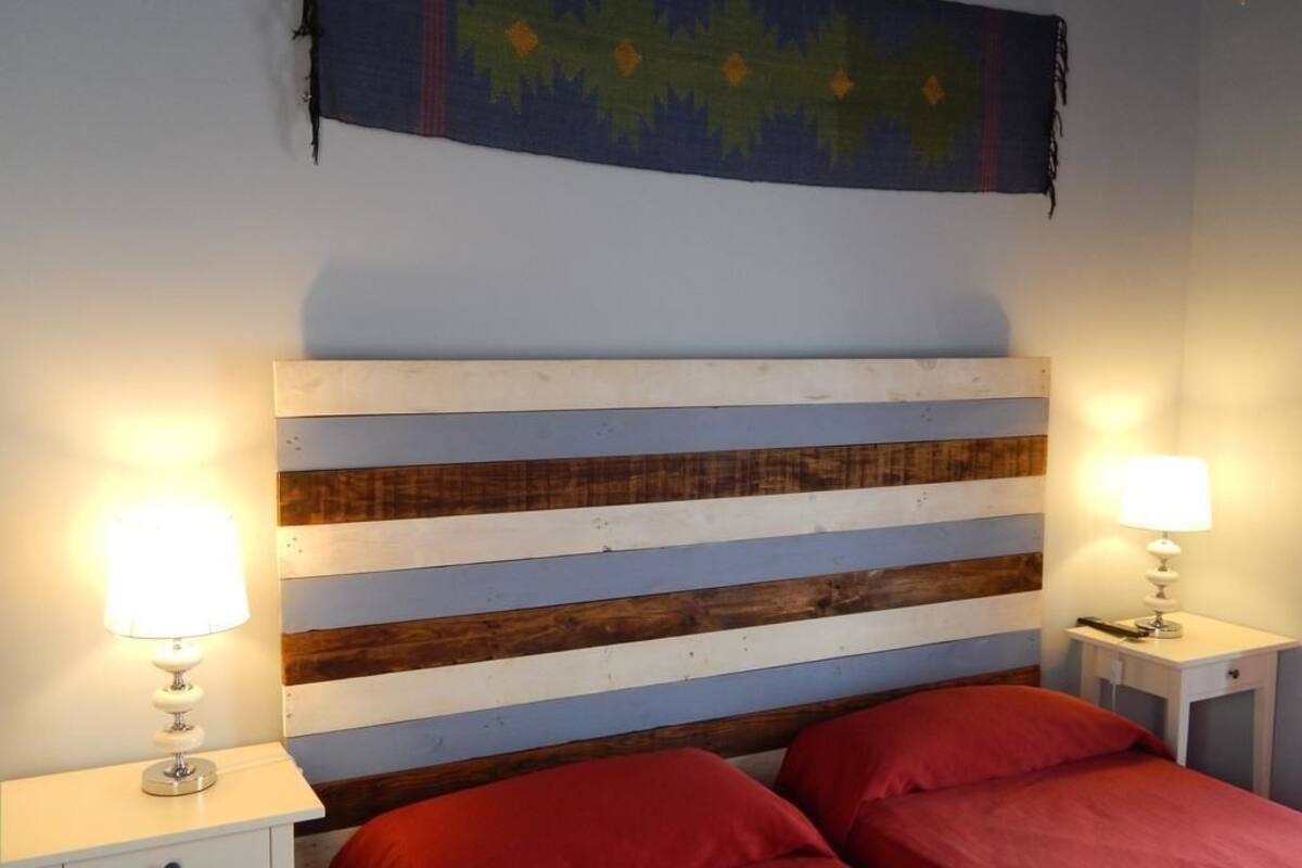 A room with a striped wooden headboard, two bedside tables with lamps, and two red beds.