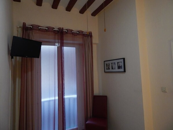 Una habitación con televisión montada en la pared, una ventana con cortinas, un cuadro enmarcado y una silla roja.