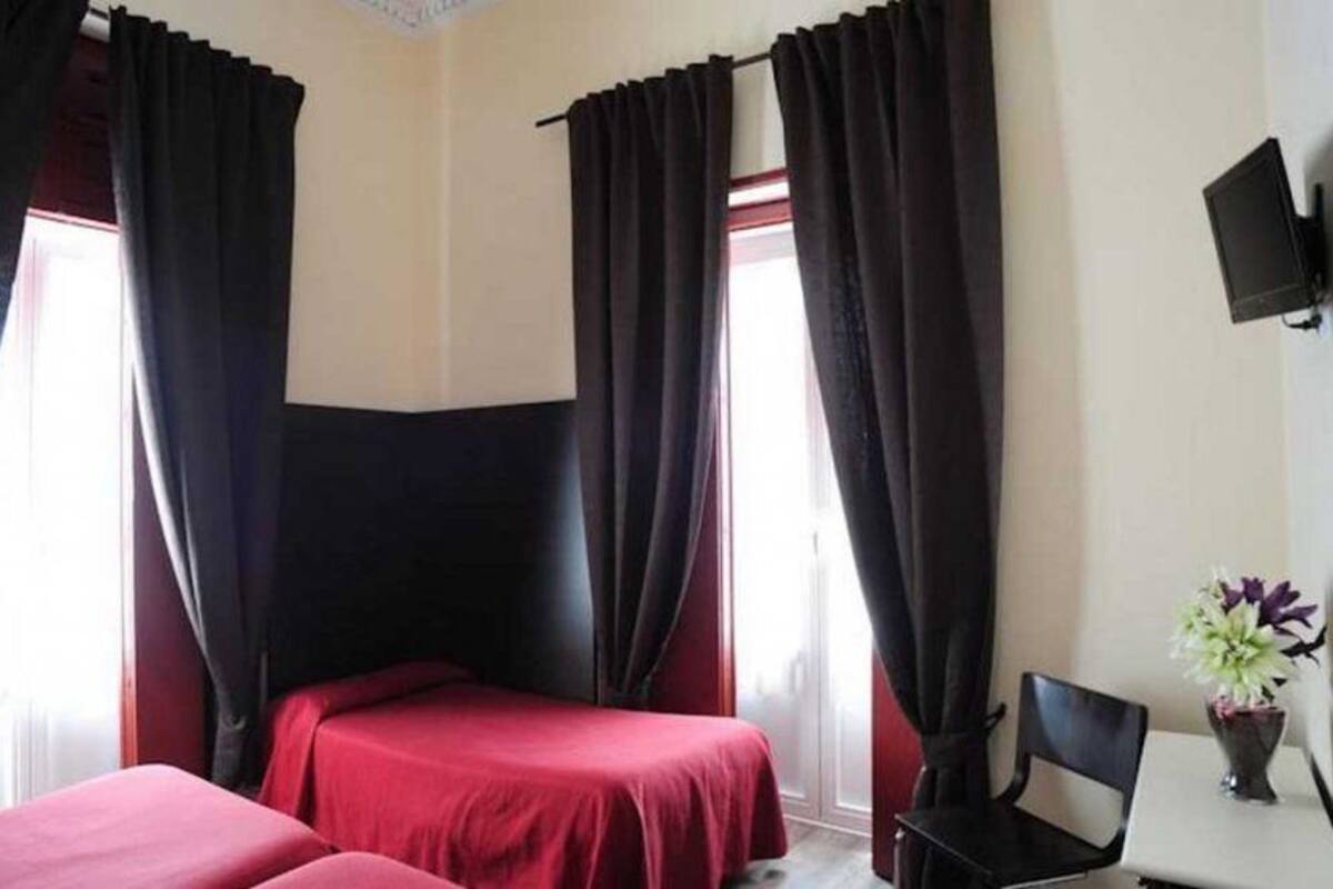 A hotel room with two beds, black curtains, a small table with a vase, and a TV on the wall.