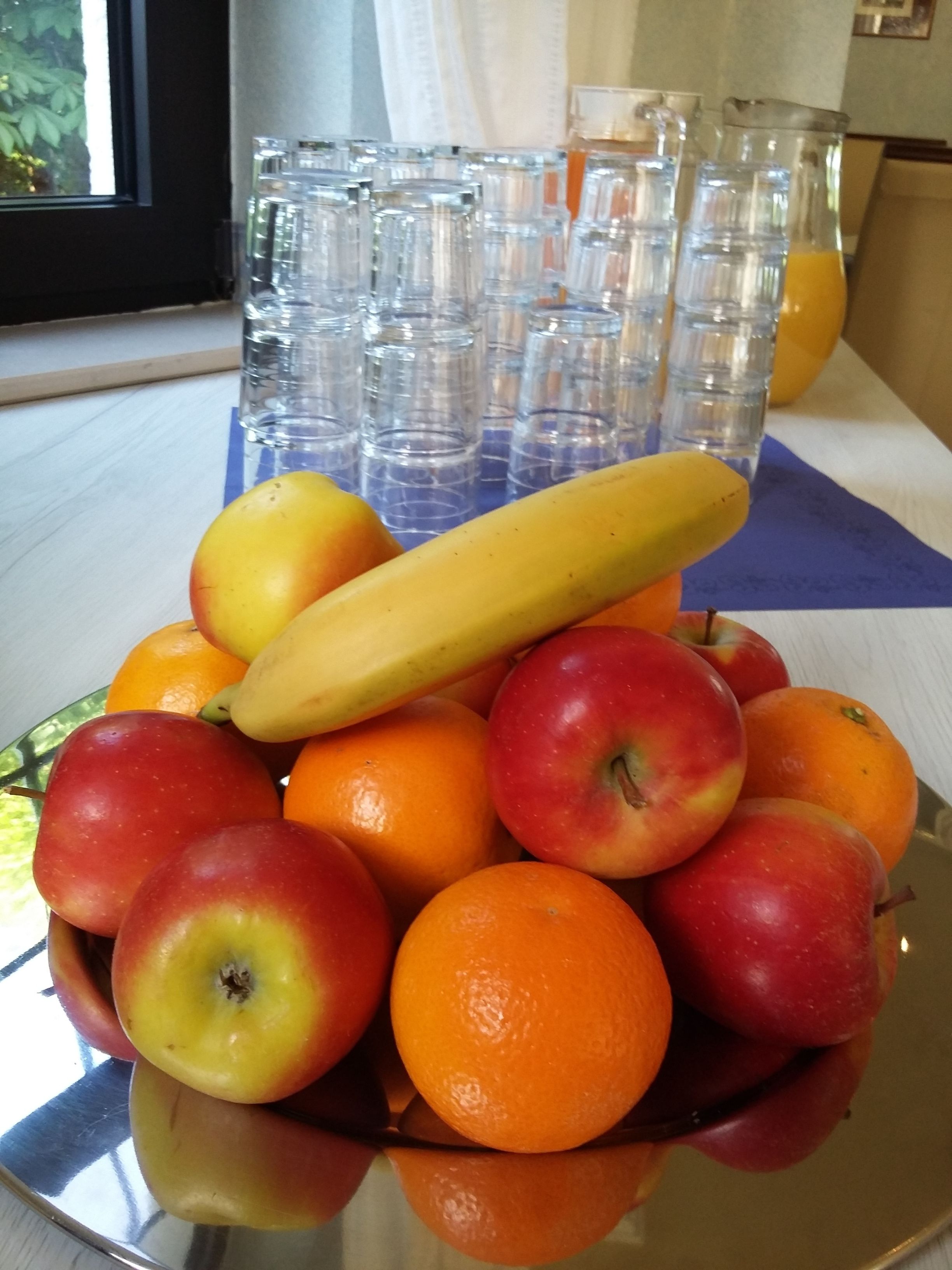 Eine Obstschale mit Äpfeln, Orangen und einer Banane auf einer spiegelnden Oberfläche.