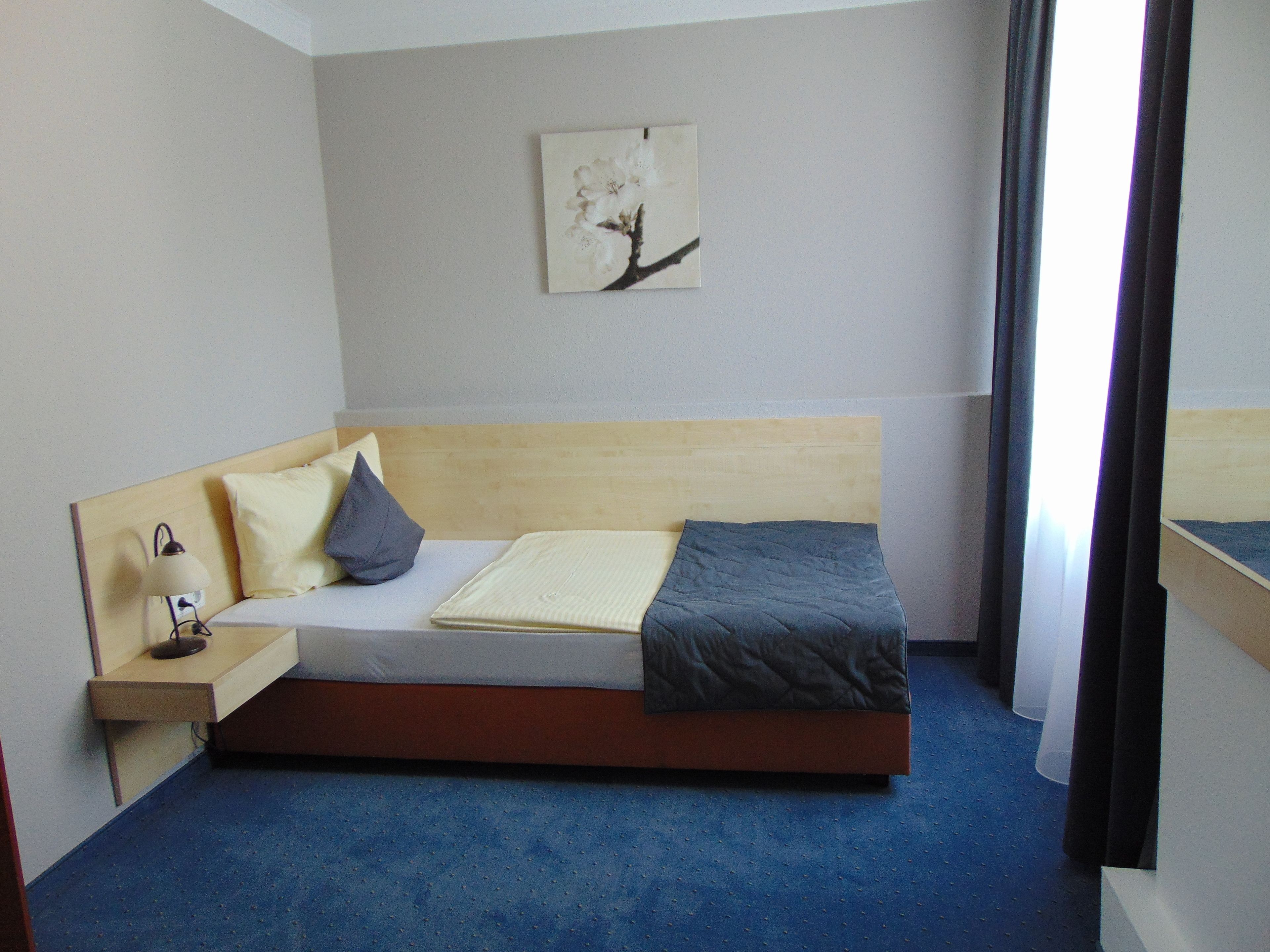 A single bed with a wooden headboard, a bedside table with a lamp, a pillow, a blanket, and a wall-mounted picture.