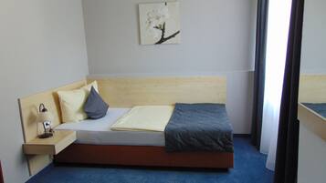 A single bed with a wooden headboard, a bedside table with a lamp, a pillow, a blanket, and a wall-mounted picture.
