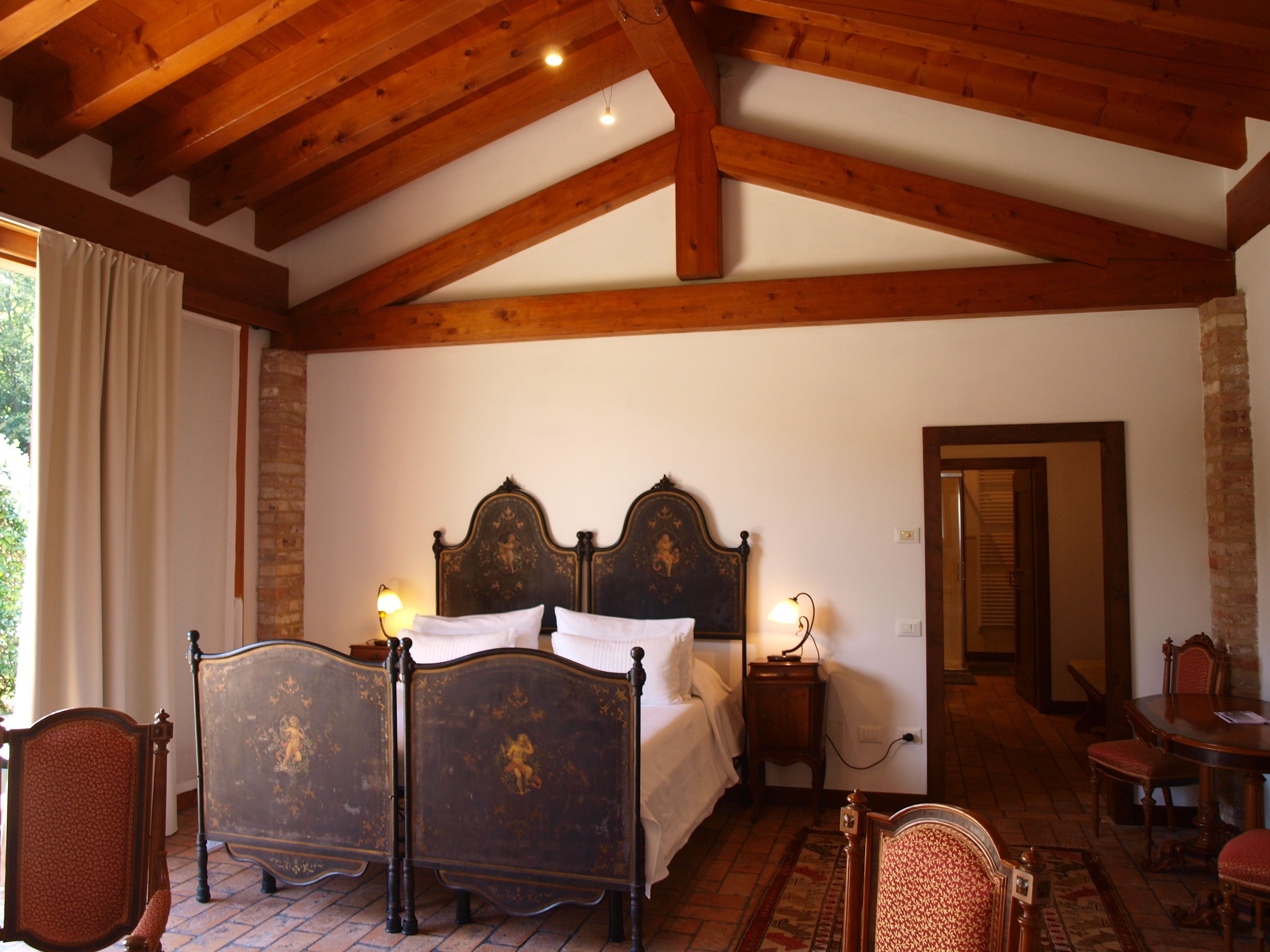 A traditional bedroom with a wooden ceiling, a large bed with ornate headboard, a bedside table with a lamp, and a view of a staircase leading to another room.